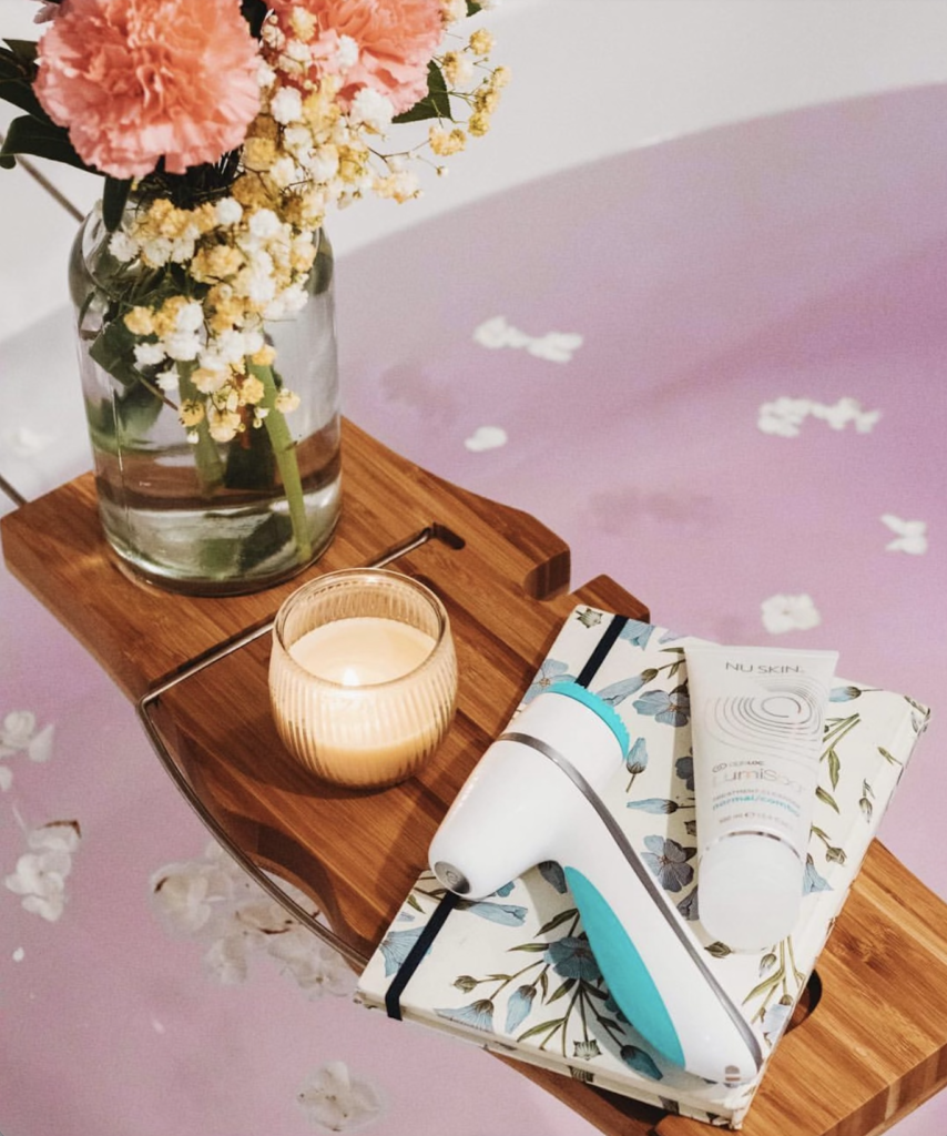A tray extends over a bathtub featuring a candle, a vase with flowers, a journal and facial products